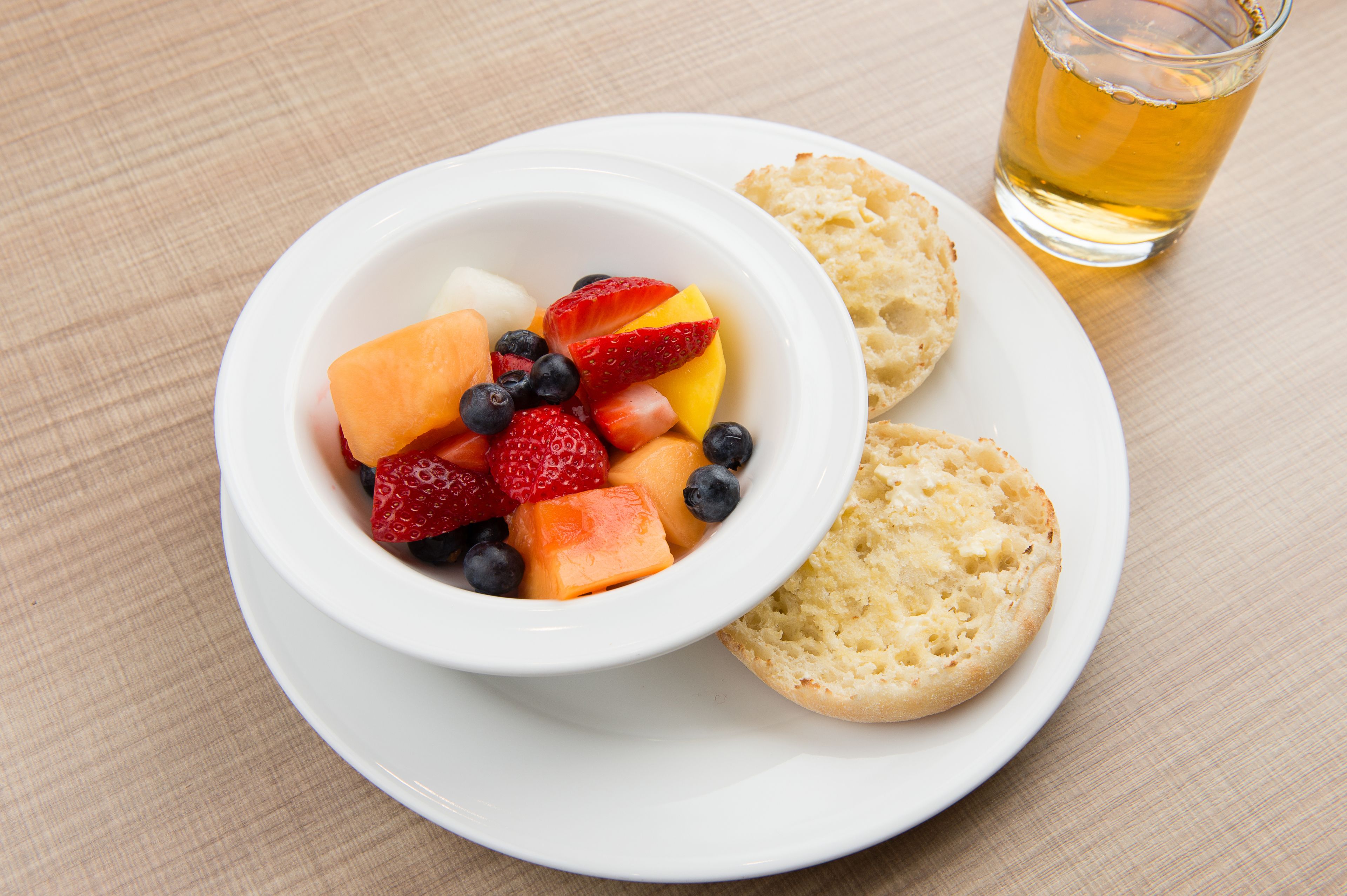 Eine weiße Schale mit gemischtem Obst auf einem weißen Teller, dazu ein Keks und eine Tasse Tee.