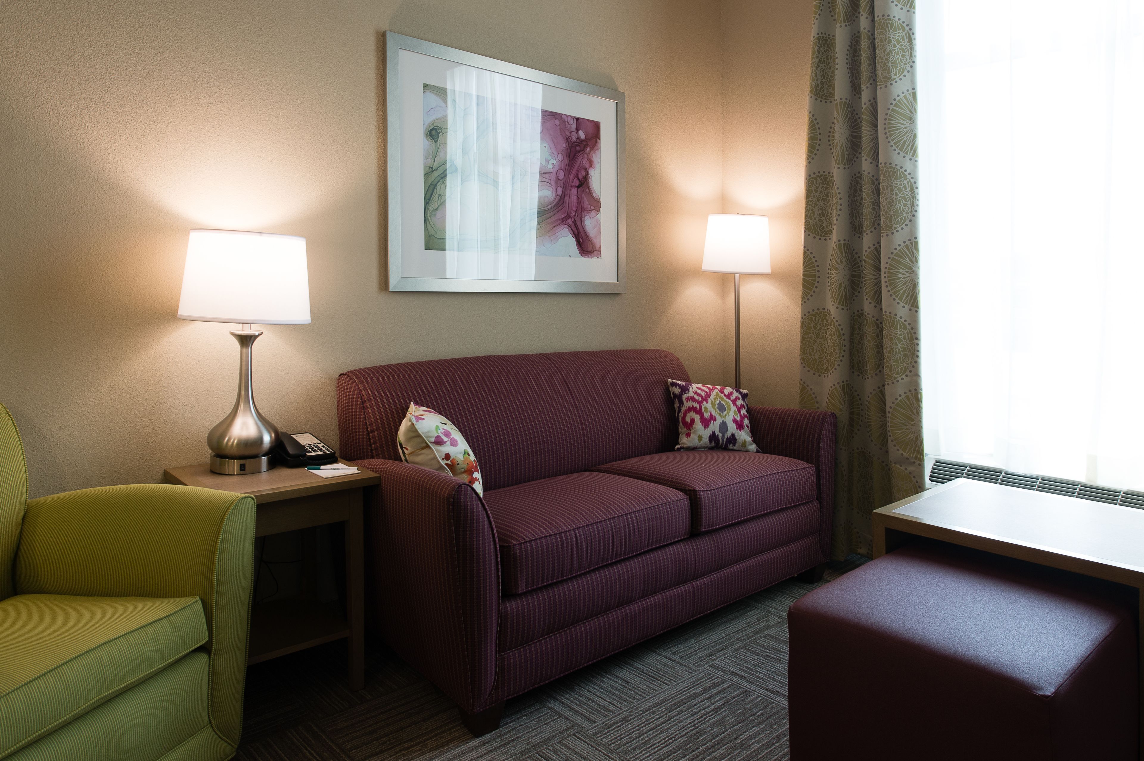 A hotel room with a green armchair, a red sofa with patterned cushions, a small table with a lamp, and a framed artwork on the wall.