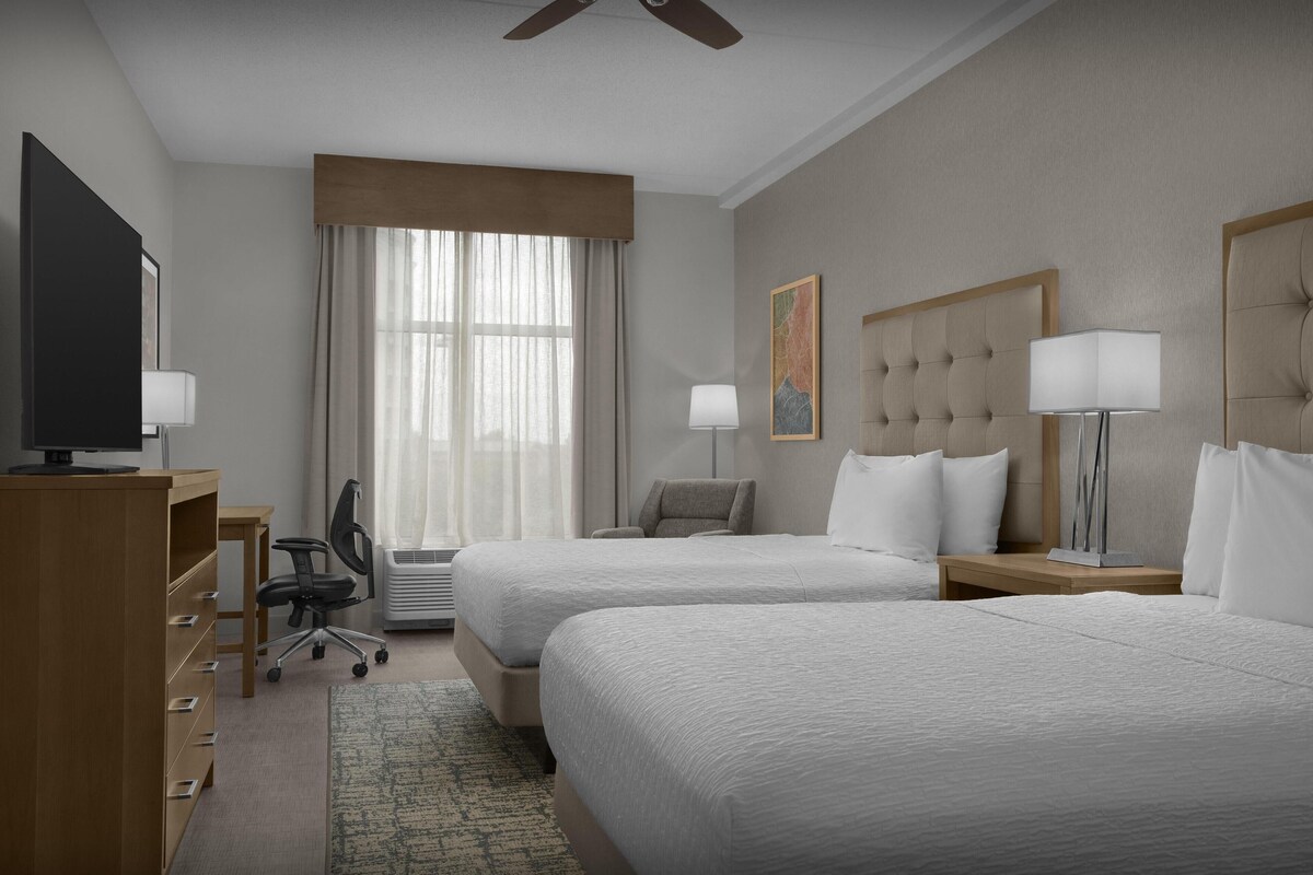 Een hotelkamer met twee bedden, een bureau, een stoel, een televisie en een groot raam met gordijnen.