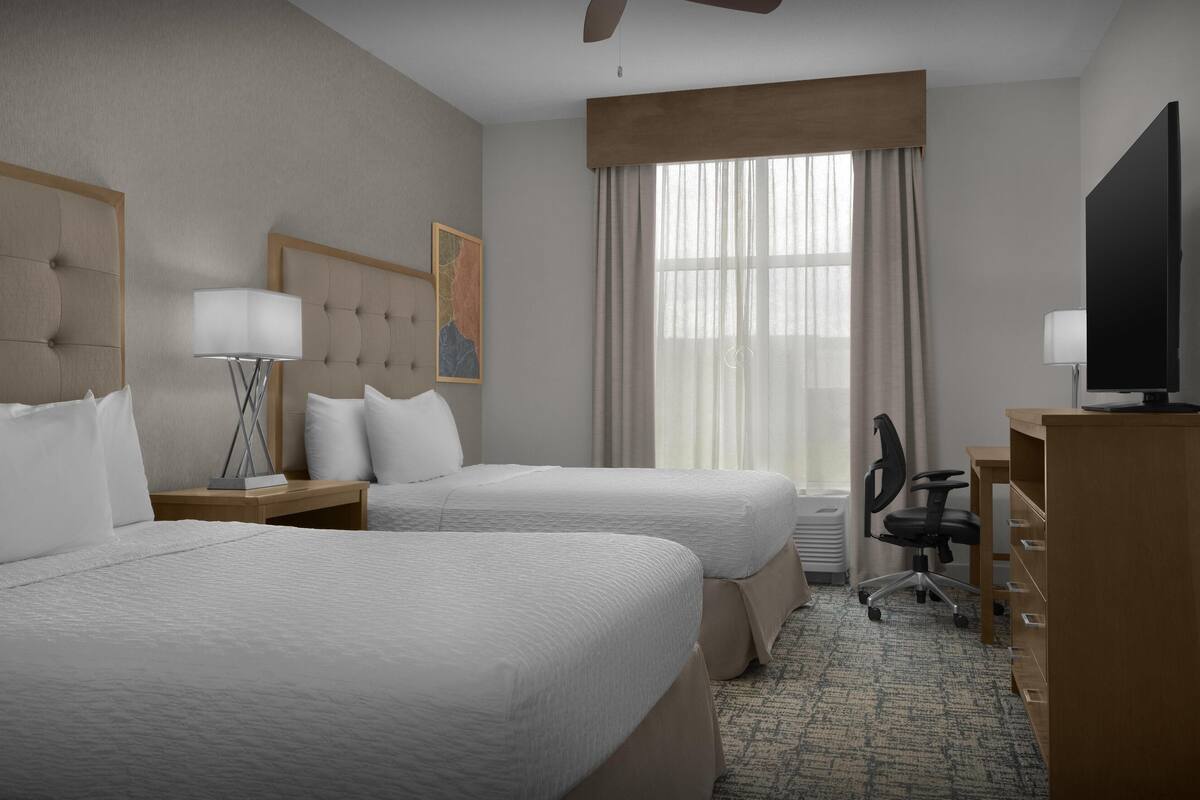A hotel room with two beds, a desk, a chair, a television, and a large window with curtains.