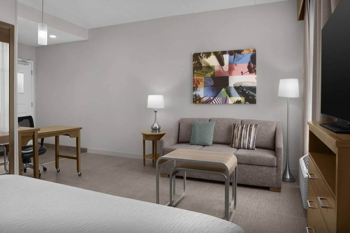 A hotel room with a bed, a desk, a sofa, a floor lamp, a TV, and a wall art piece.