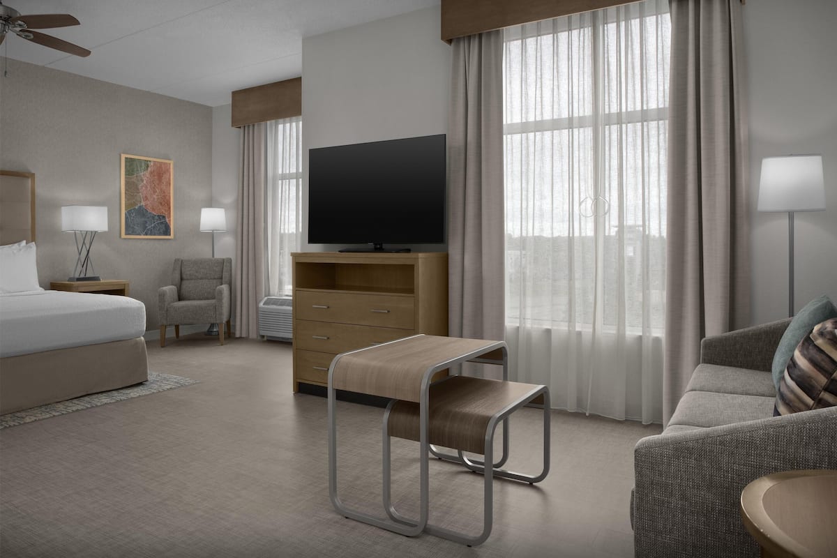 A hotel room with a bed, a TV, a desk, a chair, a sofa, and a window with curtains.
