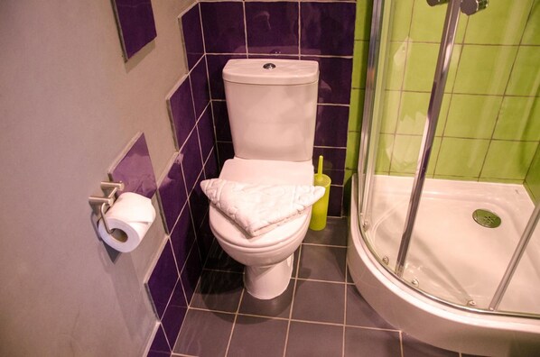 Un baño con inodoro, un rollo de papel higiénico, un cepillo amarillo de limpieza y una zona de ducha.