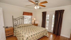 Une chambre à coucher avec un lit, une table de chevet en bois, une lampe, un ventilateur de plafond et une vue à travers une porte vitrée.