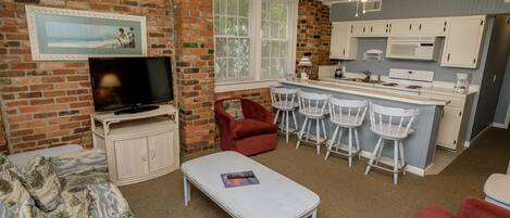A compact living space with a brick wall, a television, a sofa, a small dining area with bar stools, and a kitchenette.