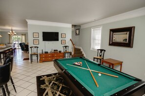 A pool table in a room with a dining area, a staircase, and a TV mounted on the wall.