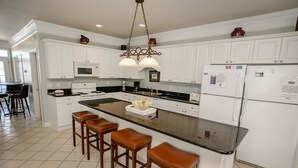 Una cocina con armarios blancos, una isla central con taburetes, microondas, refrigerador y lámpara colgante.
