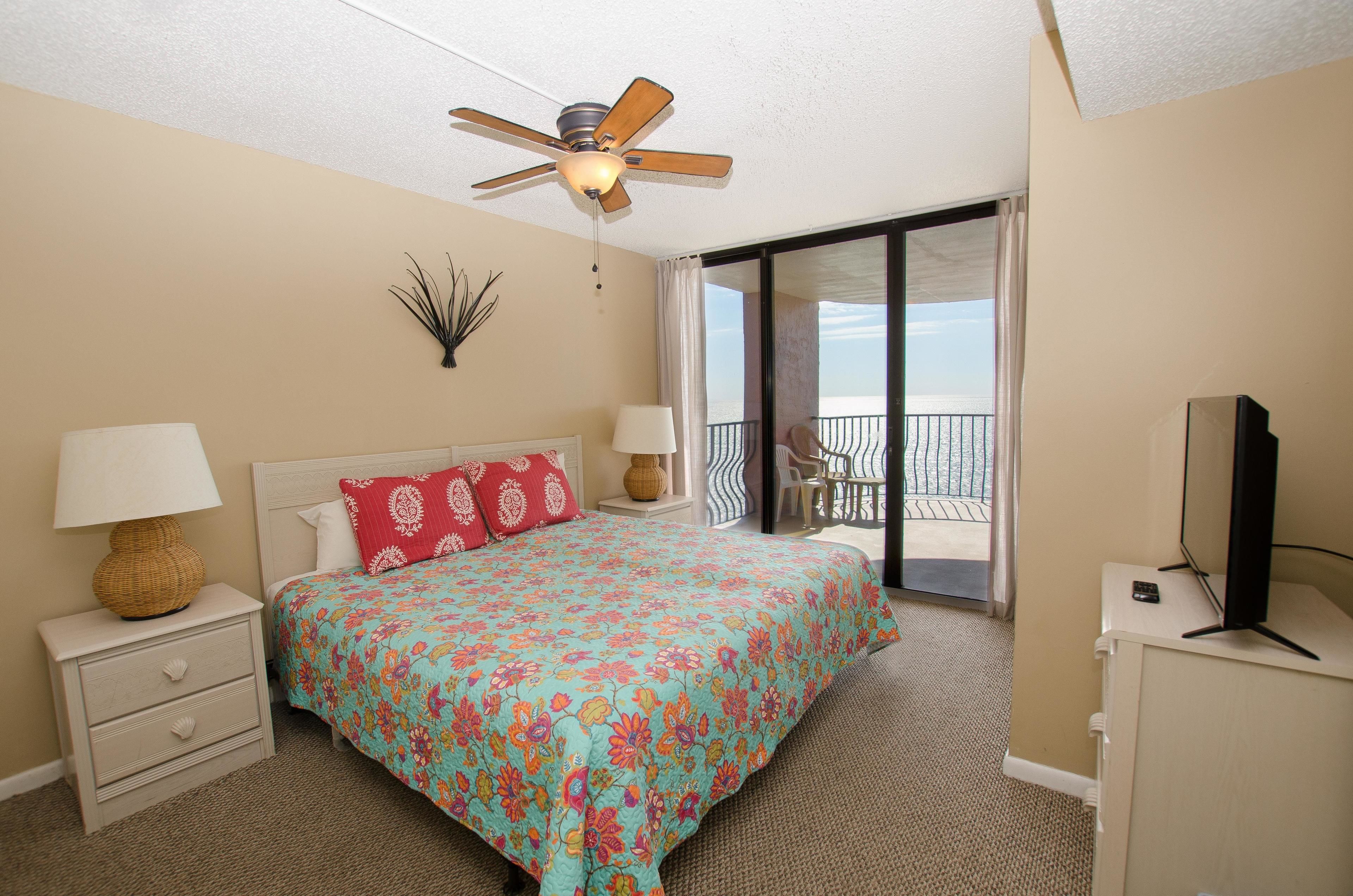 A hotel room with a bed, a ceiling fan, a TV, a nightstand, a lamp, and a view of the beach through the balcony doors.