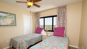 A room with two beds, each with a floral bedspread, a ceiling fan, a window with curtains, and a painting on the wall. - The Palms 1701 Villa 4 bedroom By Affordable Large Properties (Myrtle Beach)