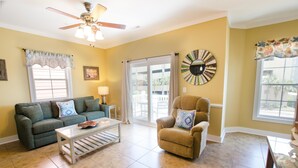 A living room with a green sofa, a beige armchair, a white coffee table, a ceiling fan, and a large window with curtains. - South Beach Cottages 4 bedroom By Affordable Large Properties (Myrtle Beach)