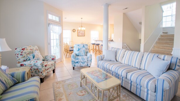 A spacious living room with a striped sofa, floral armchair, and a glass coffee table. A dining area with a table and chairs is visible in the background.