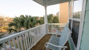 A covered porch with two chairs and a view of palm trees and a building in the background.