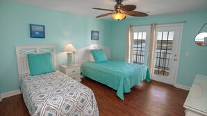 A bedroom with two beds, a ceiling fan, a view of the beach through a door, and a white dresser.