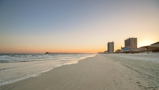 A beach with a pier extending into the ocean, buildings along the shore, and a clear sky.