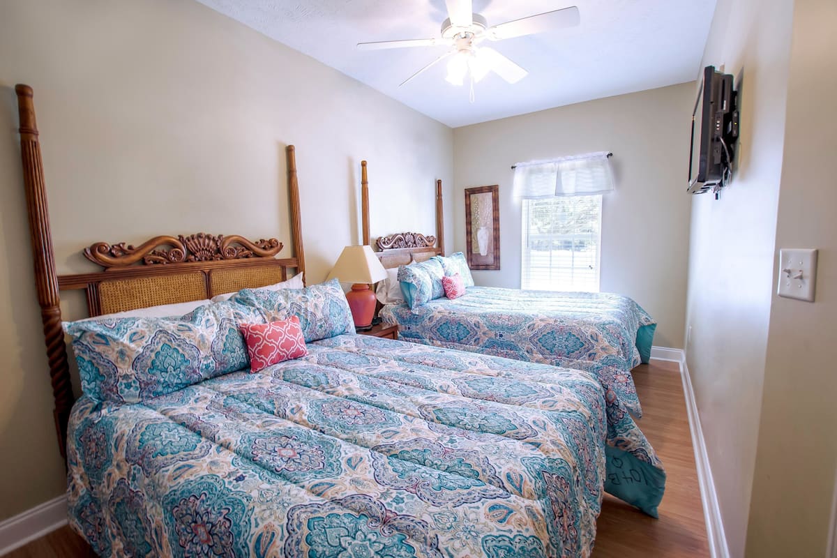 A bedroom with two beds, a ceiling fan, a window with a curtain, and a mounted TV.