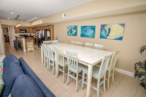 A spacious dining area with a white table and chairs, a kitchen in the background, and three paintings on the wall.