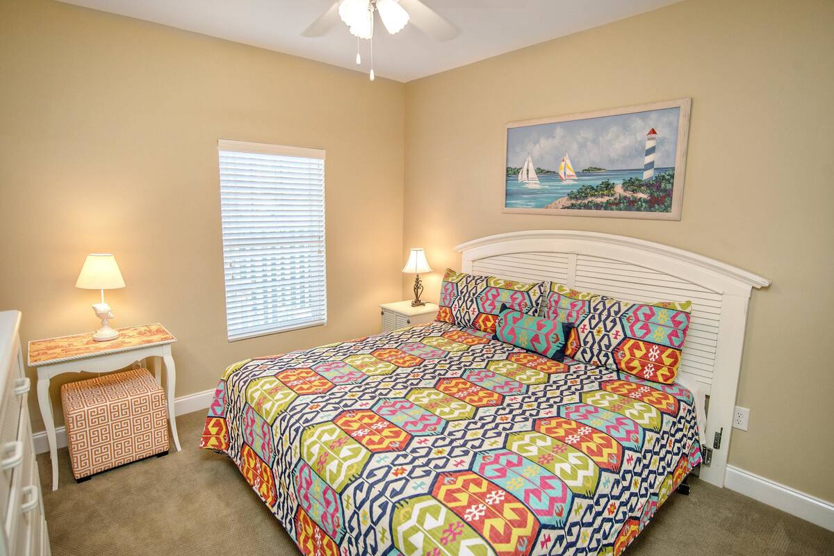A bedroom with a bed, two bedside tables with lamps, a window with blinds, a painting of a lighthouse, and a patterned ottoman.