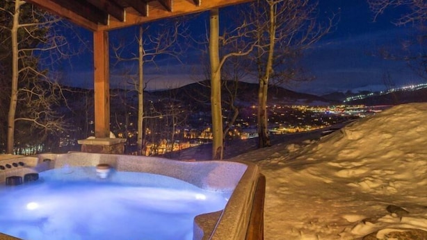 Un bain à remous à l’extérieur en pierre, entouré de neige et d’arbres, offrant une vue sur le paysage urbain illuminé la nuit.