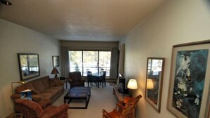 A living room with a sofa, armchair, coffee table, and a dining table with chairs. There are framed pictures on the walls and a lamp on the table.