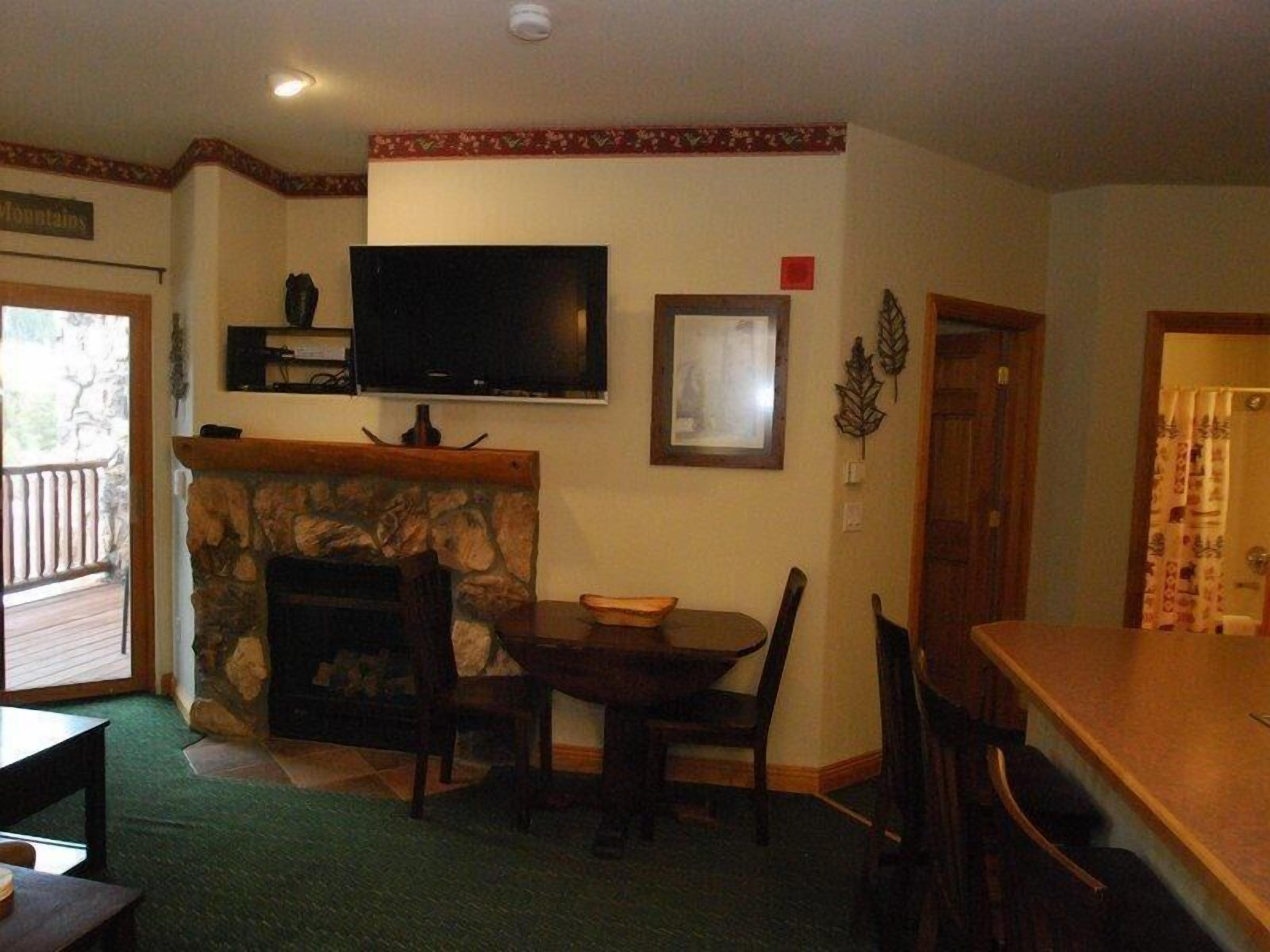 A living room with a stone fireplace, a television mounted above it, a dining table with chairs, and a wooden door.
