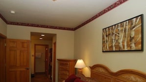 Une chambre d’hôtel avec une tête de lit en bois, une couvre-lit à motifs, un oreiller rouge, une table de chevet avec une lampe et un tableau représentant des arbres accroché au mur.