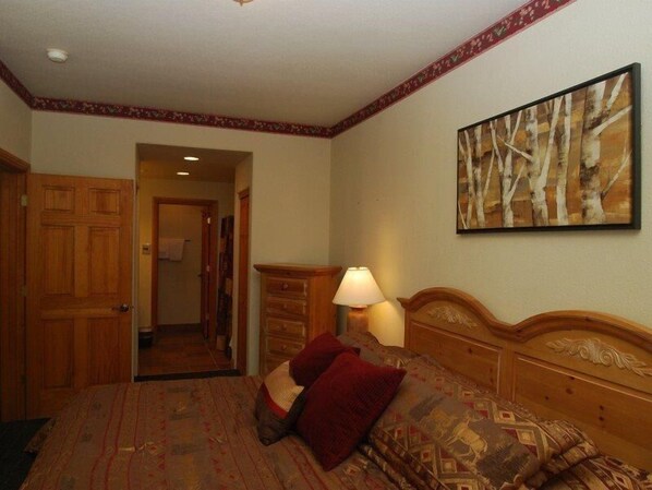 A hotel room with a wooden headboard, a patterned bedspread, a red pillow, a nightstand with a lamp, and a painting of trees on the wall.