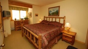 A bedroom with a wooden bed, a bedside table with a lamp, a television on a stand, and a window with curtains.