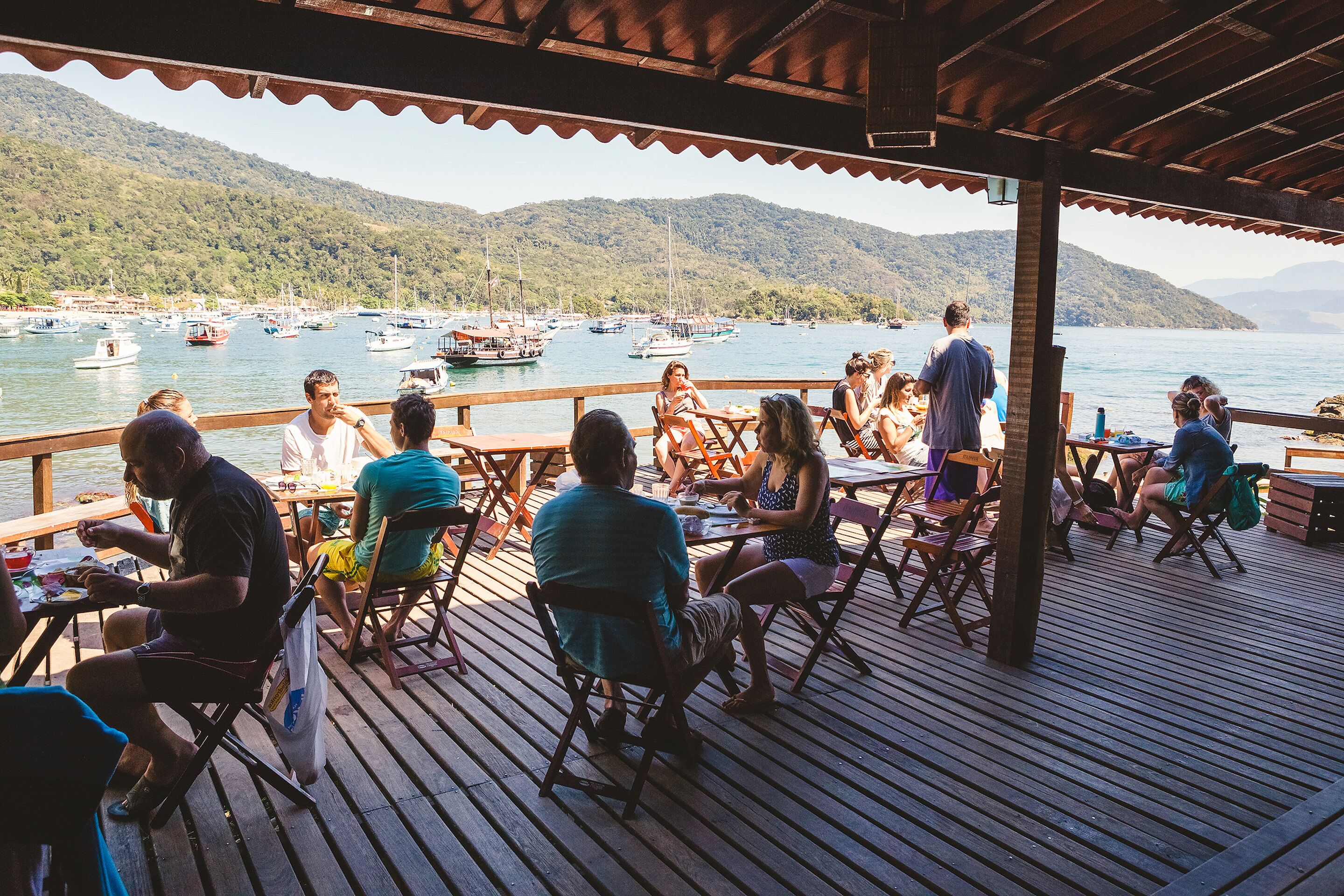 A wooden deck with tables and chairs overlooking a marina with boats and hills in the background.