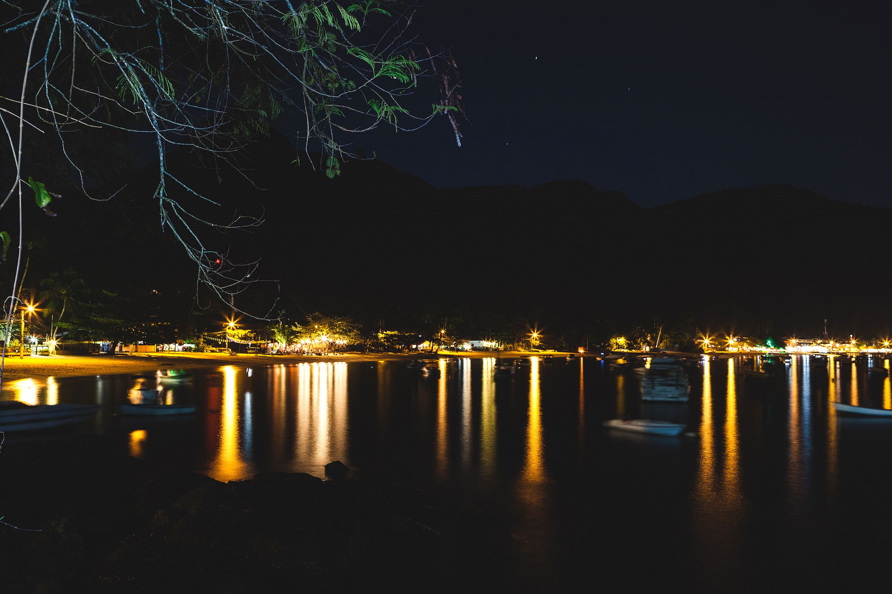 Vista nocturna de un lago tranquilo con barcos y luces distantes reflejándose en el agua.