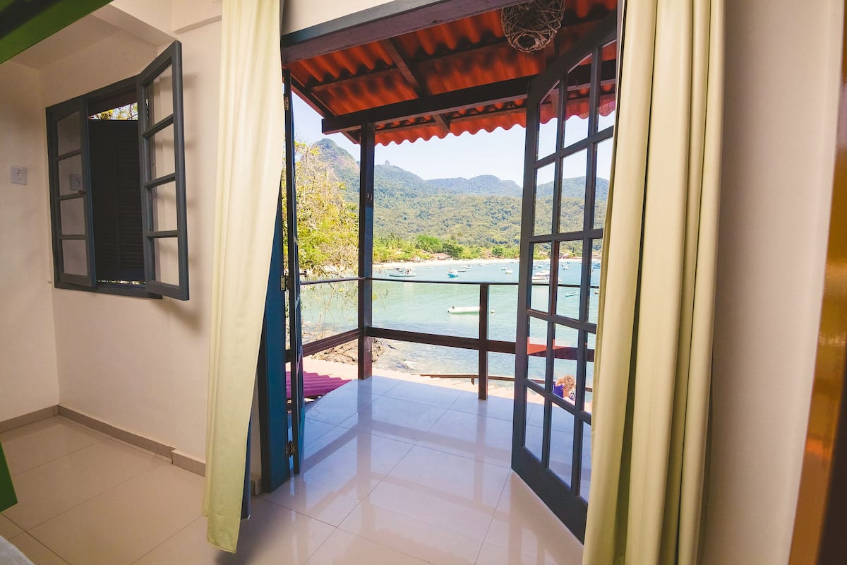 A room with a view of a beach and mountains, a window with a curtain, and a tiled floor.