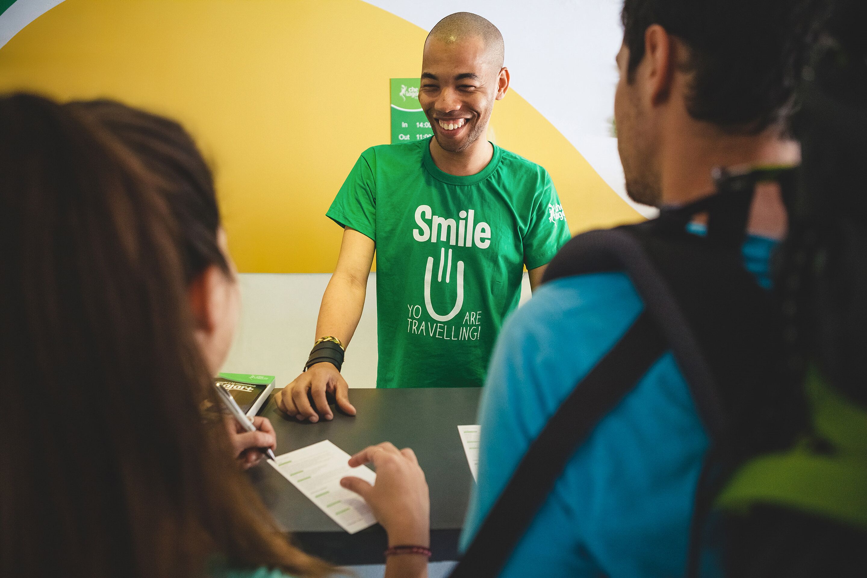 Eine Person mit grünem T-Shirt mit dem Schriftzug 'Smile Yo are Travelling!' steht an einem Schalter mit Unterlagen.