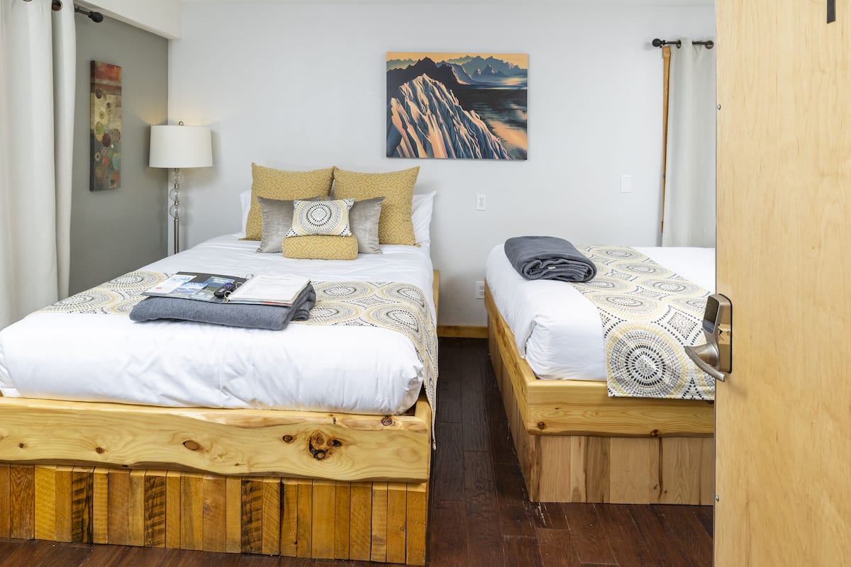 A room with two wooden beds, each with a patterned bedspread and pillows. There is a lamp on the left side, a painting on the wall, and a wooden door on the right.