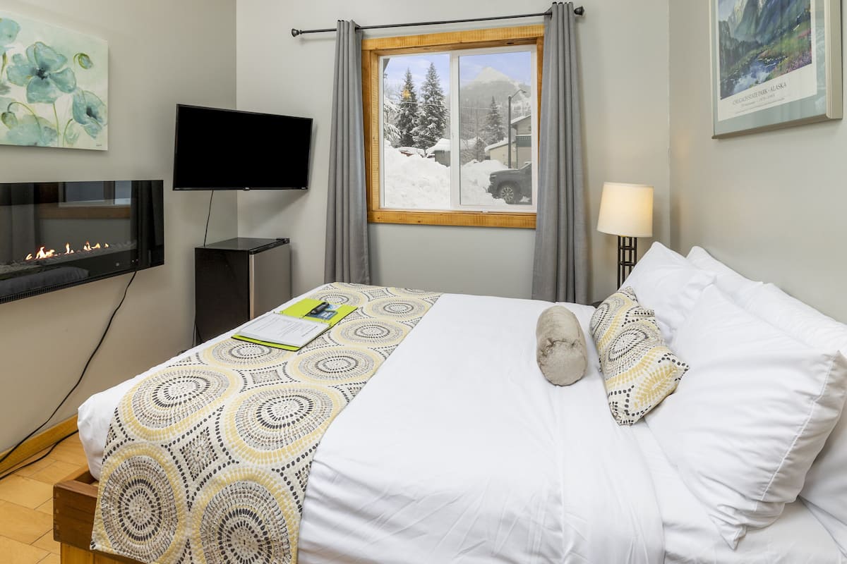 A hotel room with a bed, a flat-screen TV mounted on the wall, a fireplace, a window with a view of snow-covered trees and mountains, and a lamp on the bedside table.
