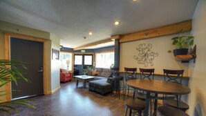 A cozy living area with a sectional sofa, a round wooden table, and a bar stool. There is a door leading to another room, a window with a view, and a wall-mounted shelf with a plant. - Ski Inn (Girdwood)