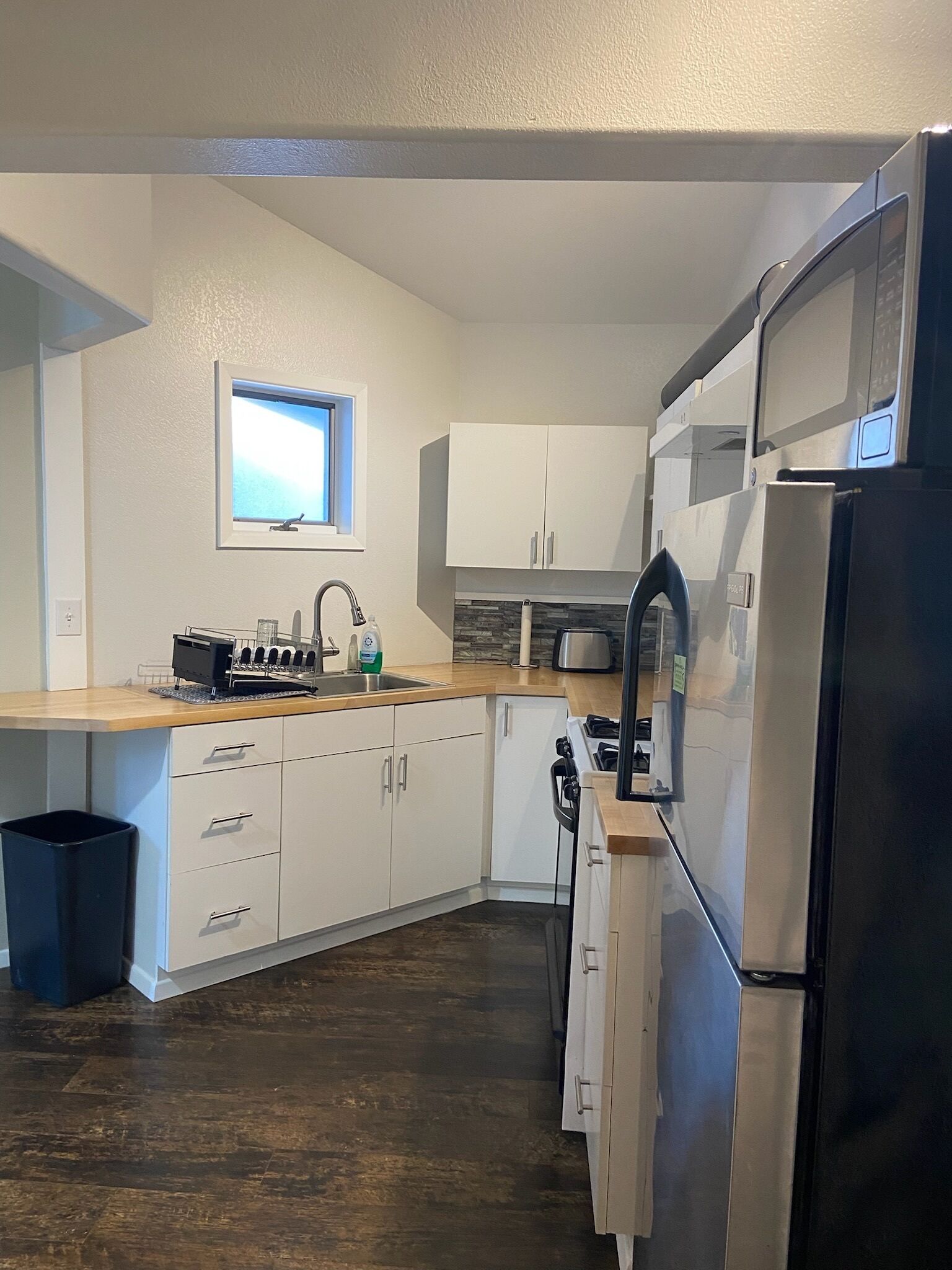A compact kitchen with white cabinets, a stainless steel refrigerator, a window, and a wooden countertop.