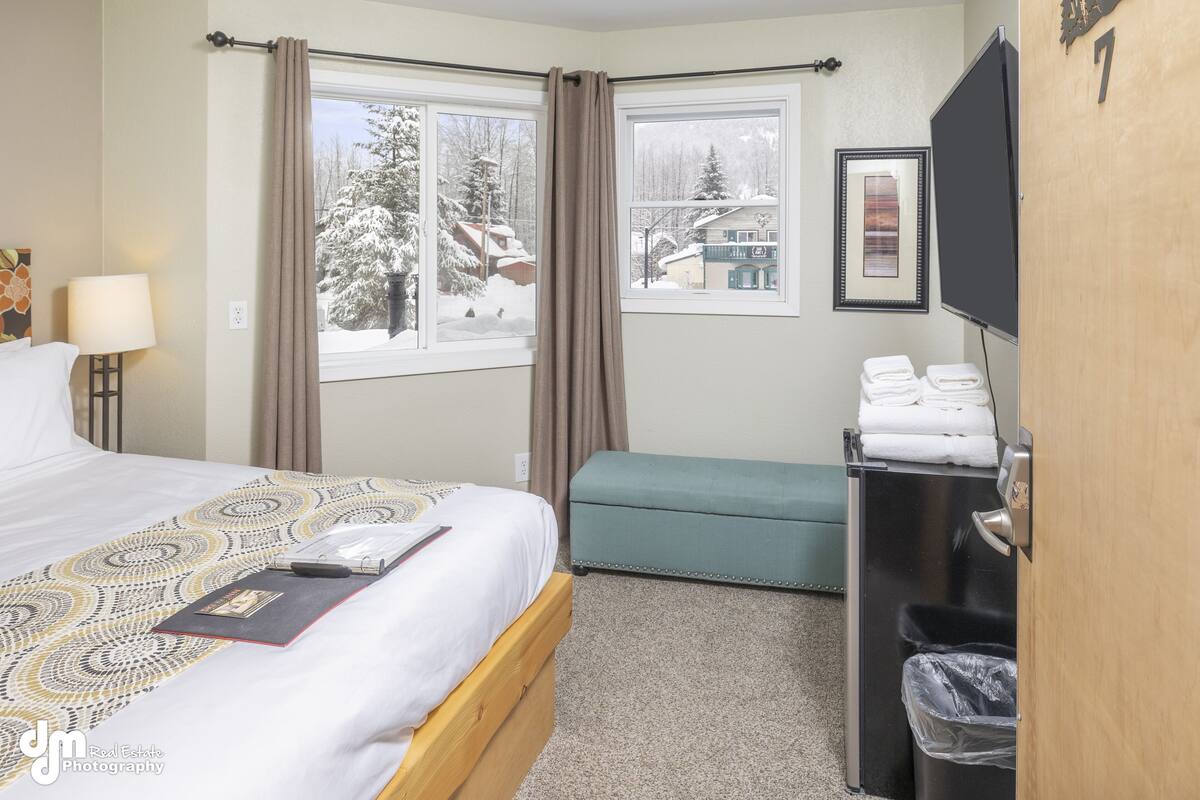 A hotel room with a bed, a bench, a TV, and a window with a view of snow-covered trees.