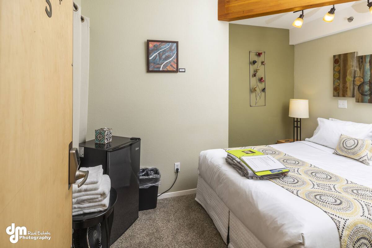 A hotel room with a bed, a desk, a trash bin, and artwork on the walls.