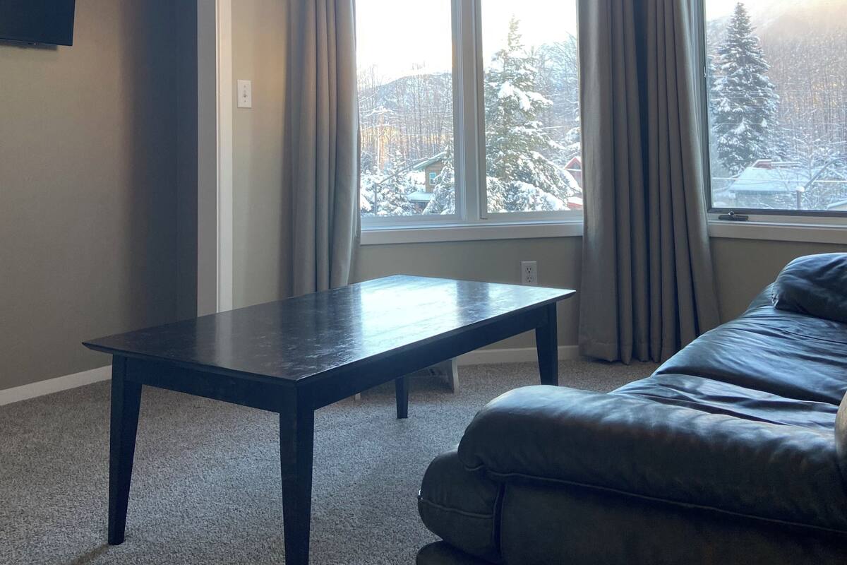 A room with a dark wooden table, a sofa, a large window with curtains, and a television on the wall.