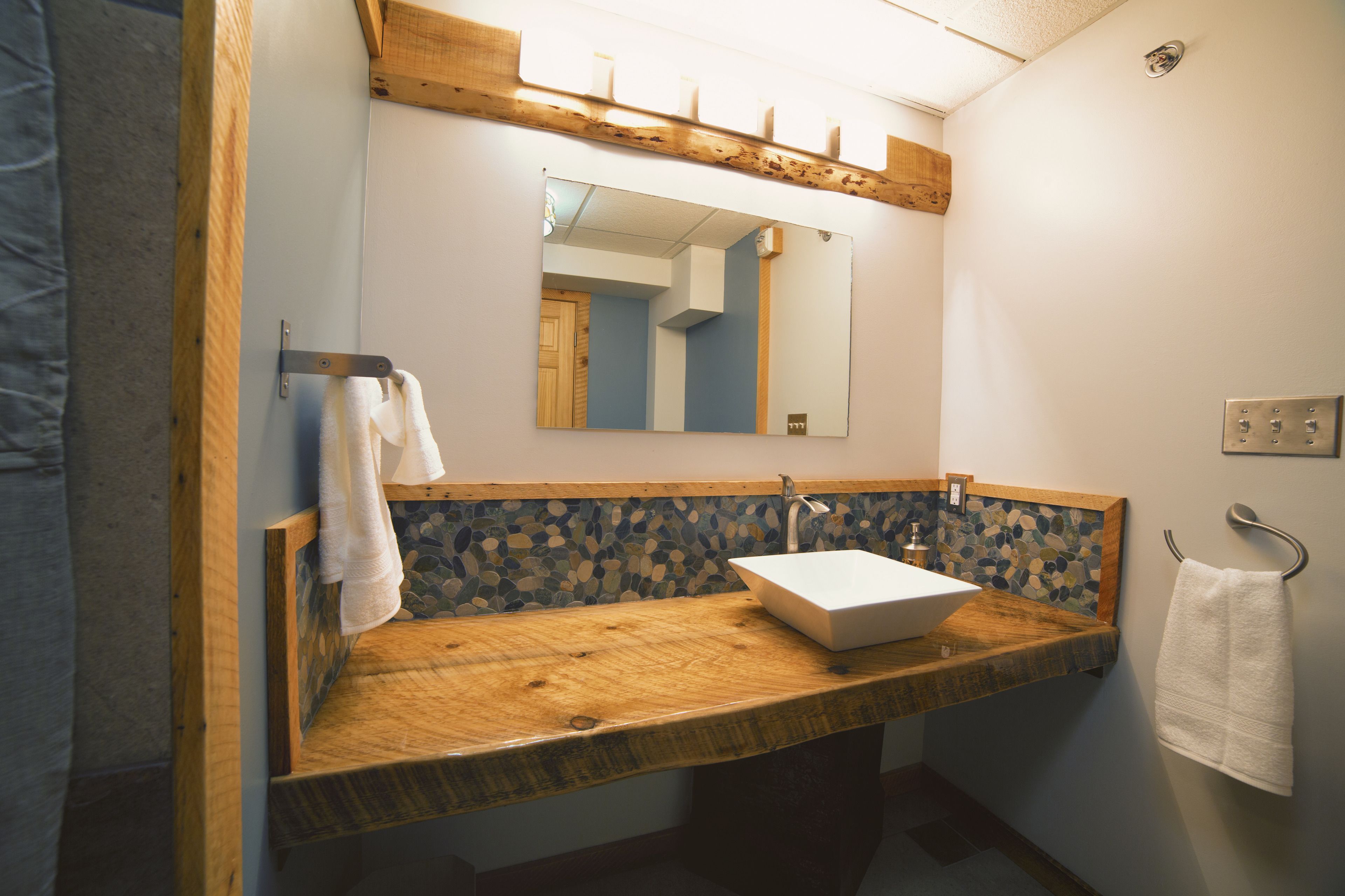 Un baño con tocador de madera, lavamanos blanco, espejo y salpicadero con mosaicos.