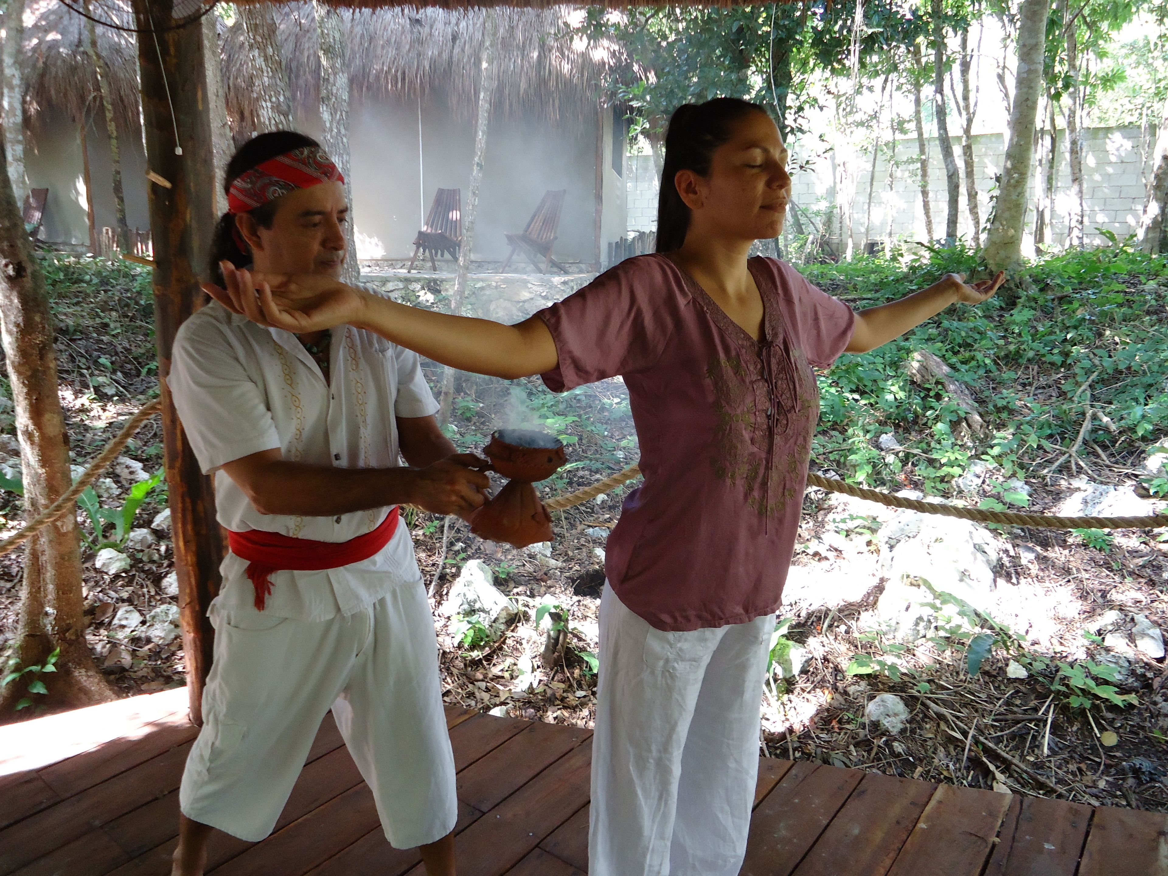 Two people practicing yoga outdoors on a wooden deck surrounded by trees.