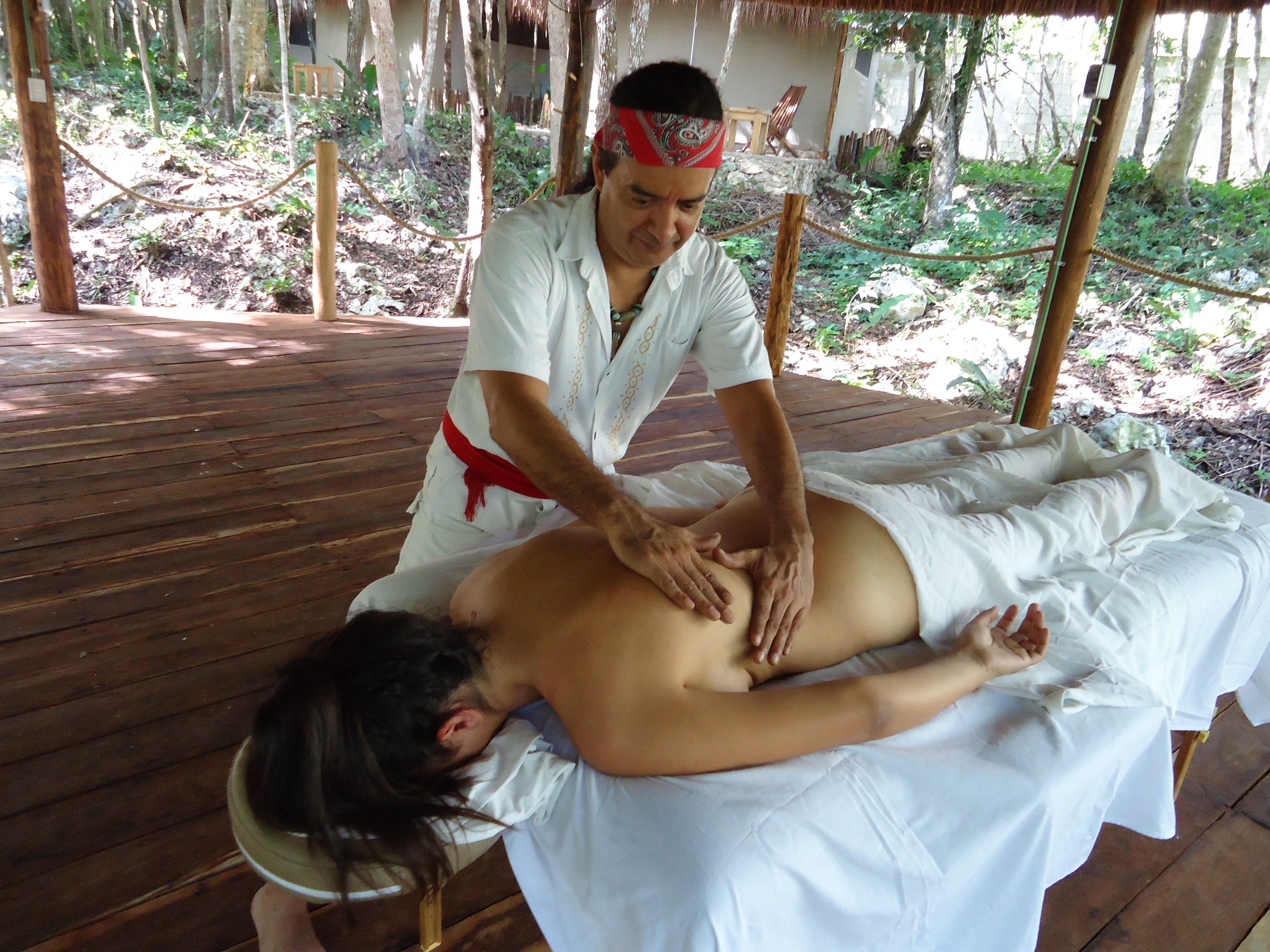 A massage therapy session in a wooden room with a thatched roof, surrounded by trees.