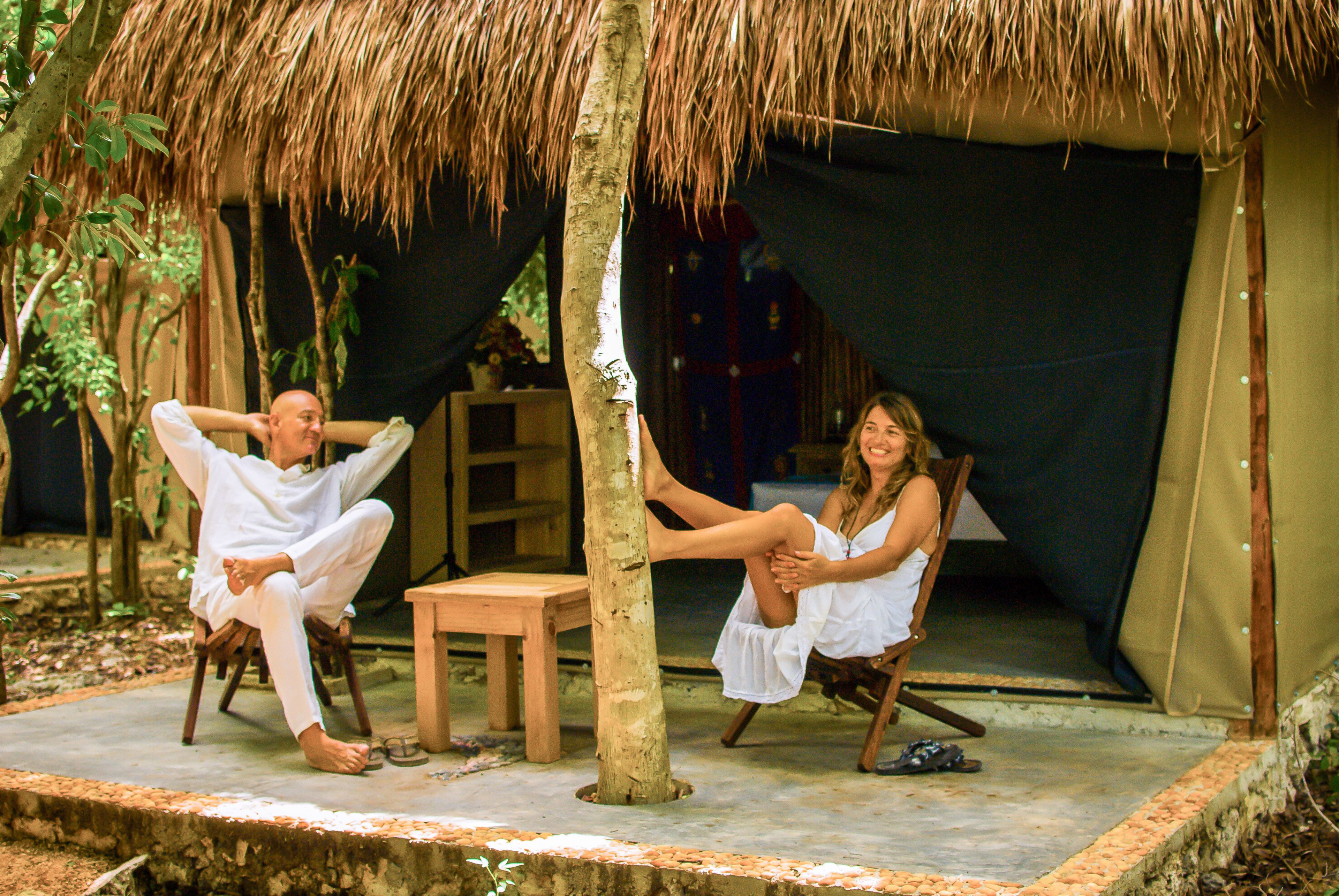 A couple relaxing under a thatched roof structure with a wooden bench and a table nearby.