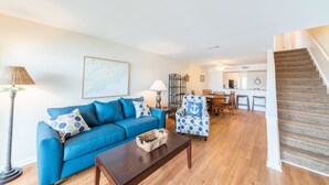 A living room with a blue sofa, a coffee table, and a patterned armchair. There is a staircase leading to an upper level. - MARINERS CAY 45 2 Bedroom Holiday Home By My Ocean Rentals (Folly Beach)