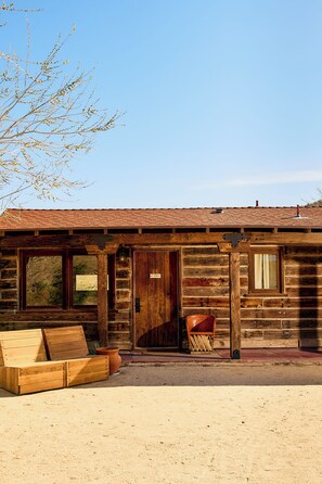 Une cabane rustique en rondins de bois, dotée d’un porche en bois, d’un banc et d’une grande fenêtre.