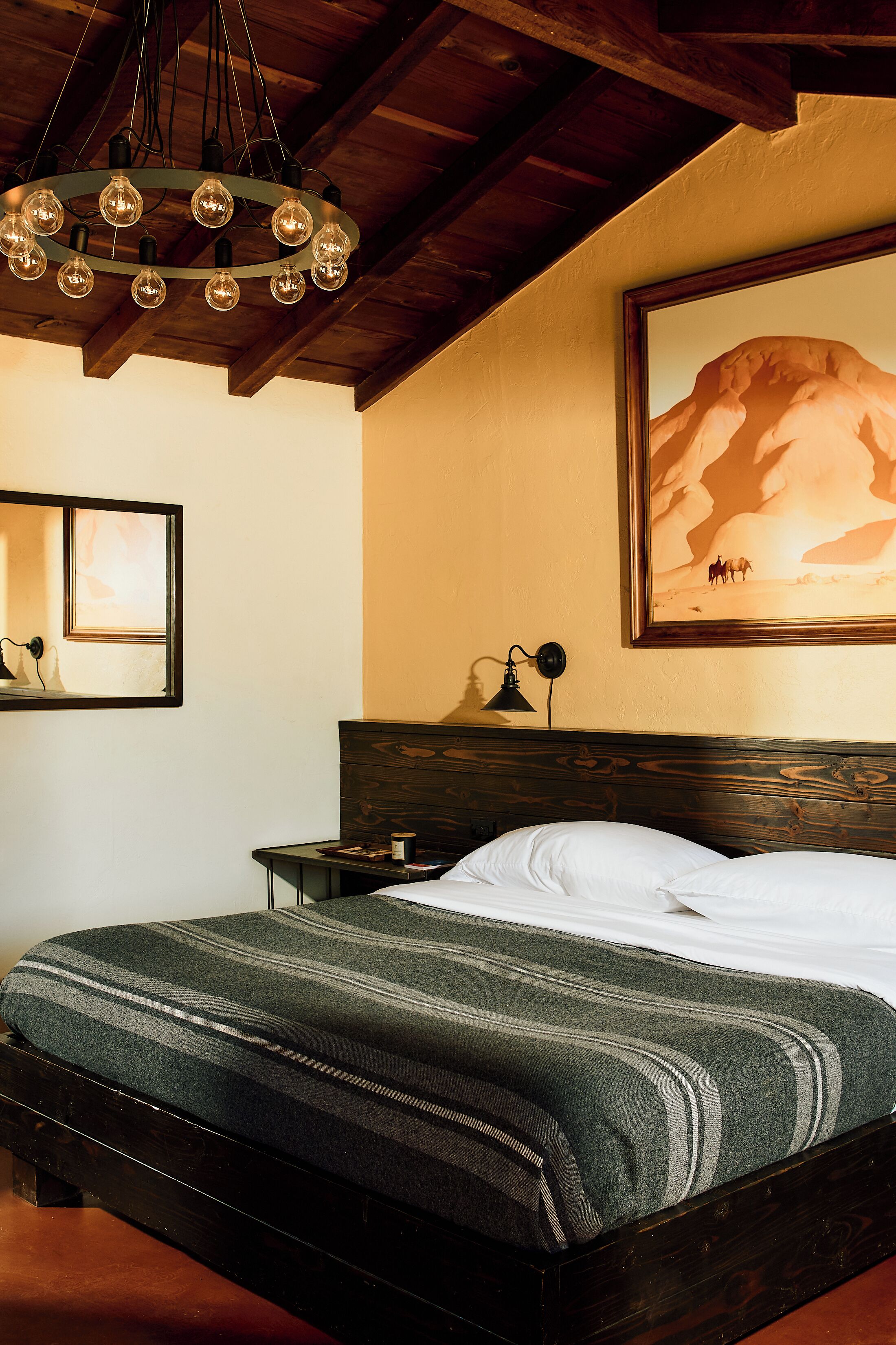 A bedroom with a wooden headboard, a bed with a striped blanket, a nightstand with a lamp, and a painting of a mountain on the wall.