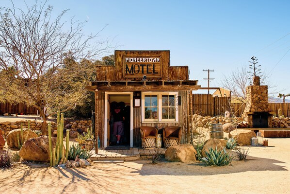 Un motel de style «ville pionnière», construit en bois, avec une façade rustique, une cheminée en pierre et un jardin désertique agrémenté de cactus et de rochers.