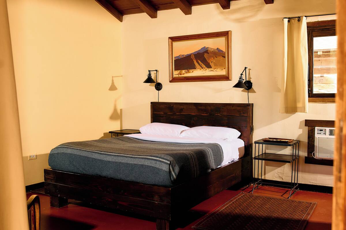 Ein hölzernes Bett mit grauer Decke, zwei Nachttischlampen, ein gerahmtes Bild einer Berglandschaft und ein Fenster mit Vorhang.