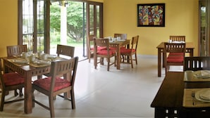 A dining area with wooden tables and chairs, a large window with a view of greenery, and a colorful abstract painting on the wall.