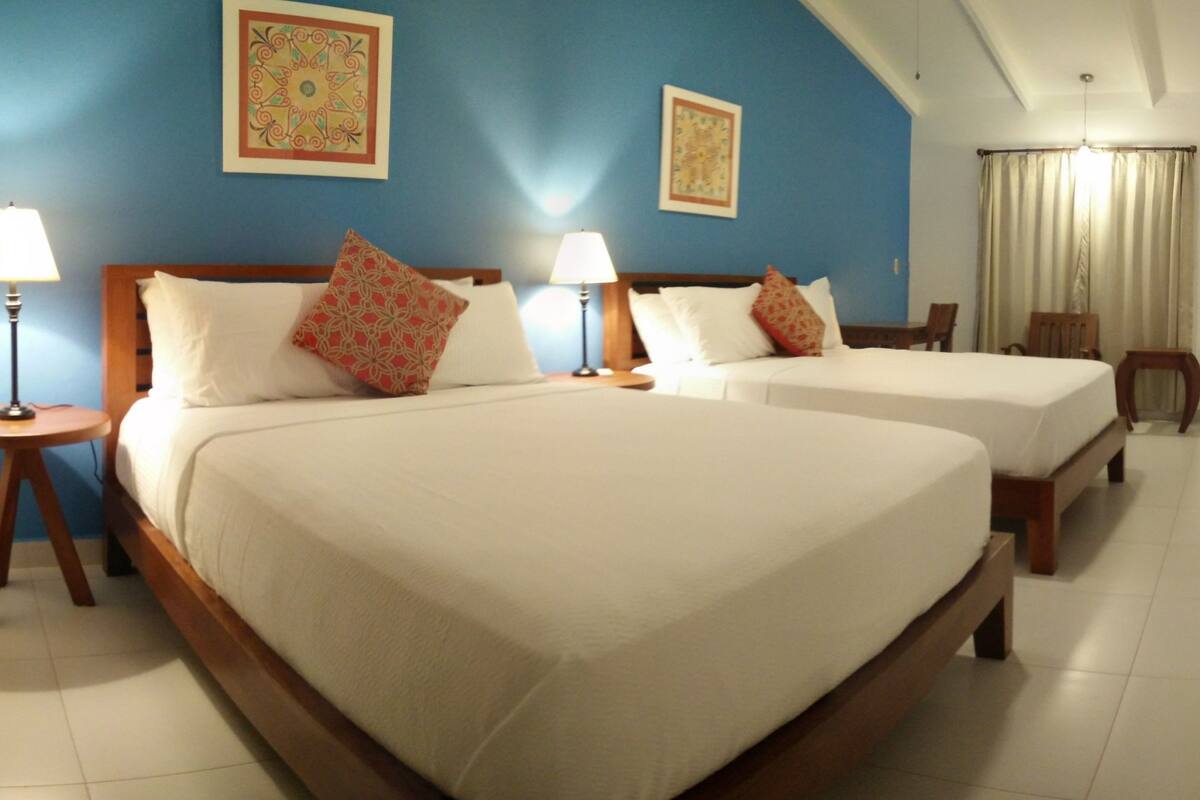 Une chambre d’hôtel avec deux lits, chacun doté d’un cadre en bois et de draps blancs. Il y a deux tables de chevet avec des lampes, et les murs sont peints en bleu.
