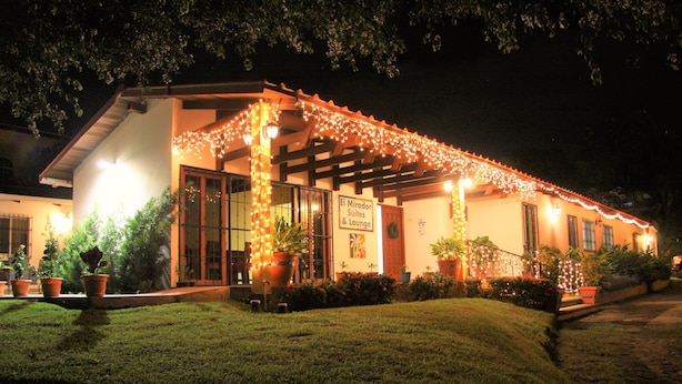 A building with a sign reading "El Mirador Suites & Lounge" at night, decorated with string lights and surrounded by potted plants.